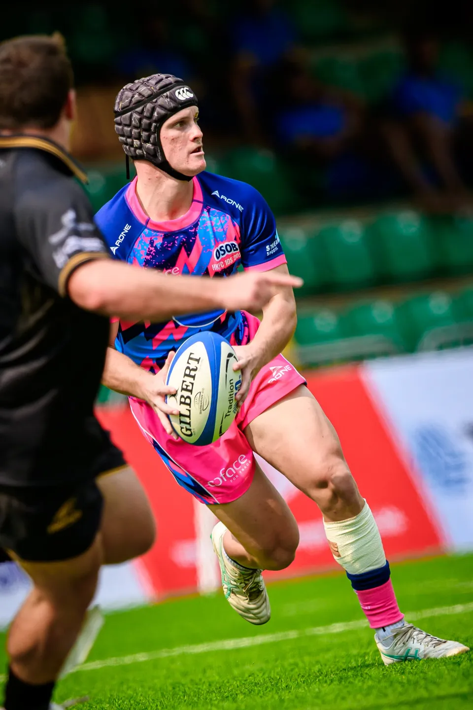 OSOB player in a scrum cap holds a Gilbert ball in both hands, looking to pass, a black sleeve in the foreground