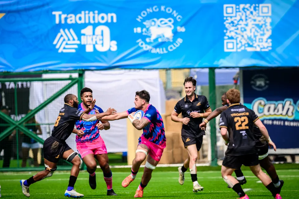 Wide view of an OSOB ball carrier running at Tropics defenders beneath a blue Tradition HKFC 10s banner