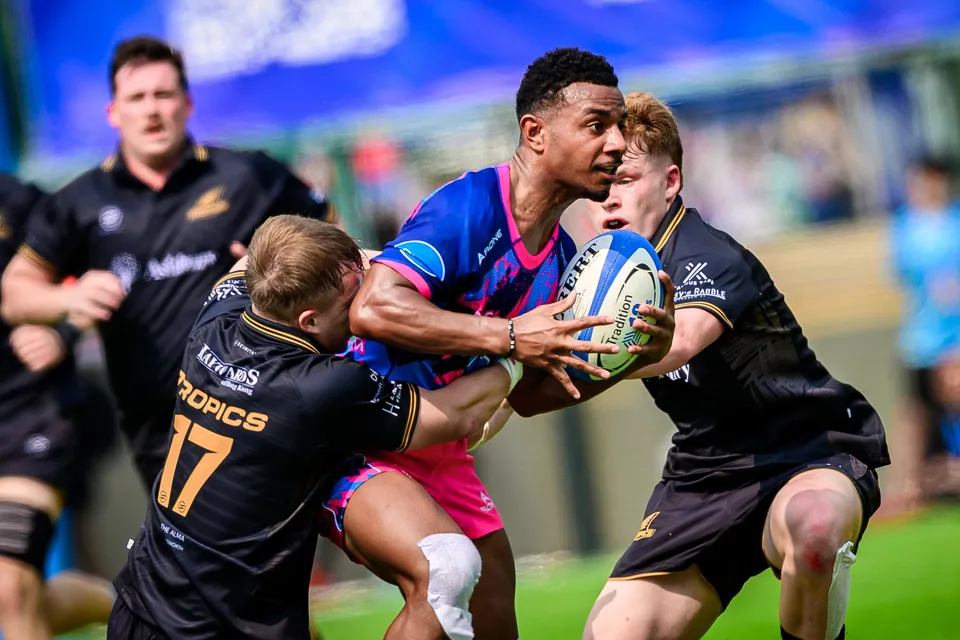 OSOB ball carrier is held up by two Tropics defenders in black, one wearing number 17, on a sunny pitch