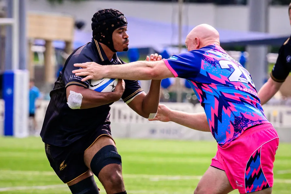 Tropics ball carrier in a scrum cap is held off by the straight arm of a bald OSOB player
