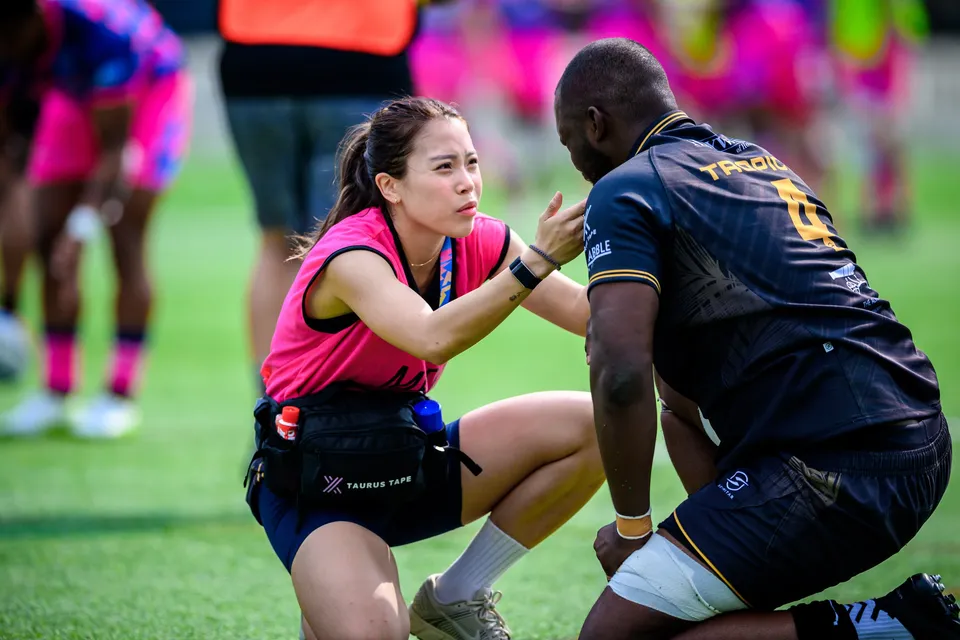 Woman in a pink top kneels beside a Tropics player in black to check on him on the pitch