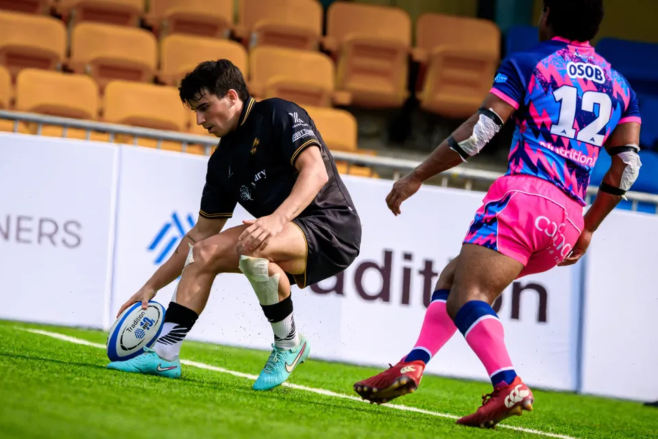 Tropics player in black dives to ground the ball by the try line as OSOB number 12 stands over him