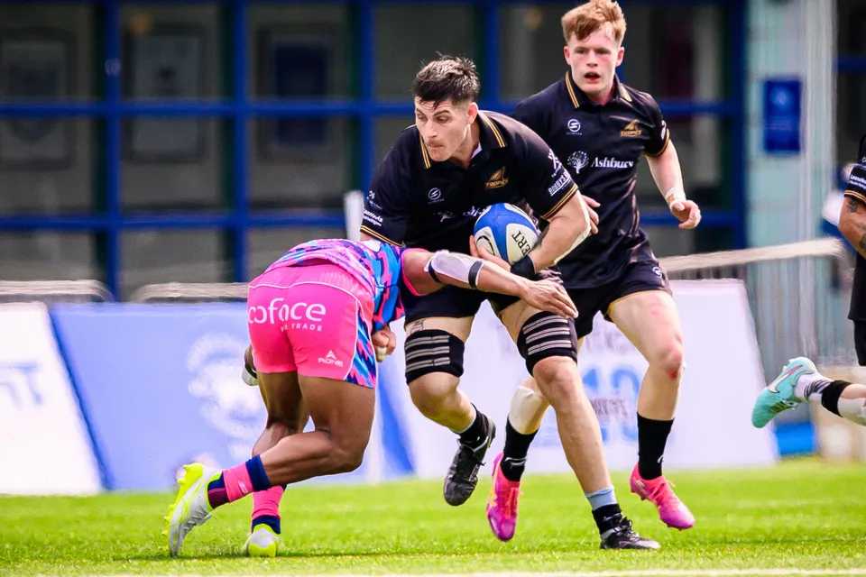 OSOB tackler brings down a black-shirted Tropics ball carrier low as a teammate in black runs in support