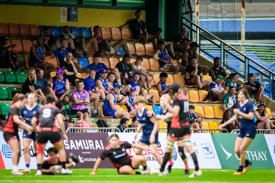 Spectators on tiered seats watch from behind blurred players in red and navy at the pitch edge, 2026 HKFC 10s
