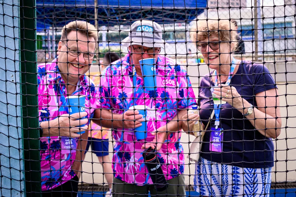 Three smiling spectators in pink floral shirts and an OSOB cap hold blue cups behind a net, 2026 HKFC 10s