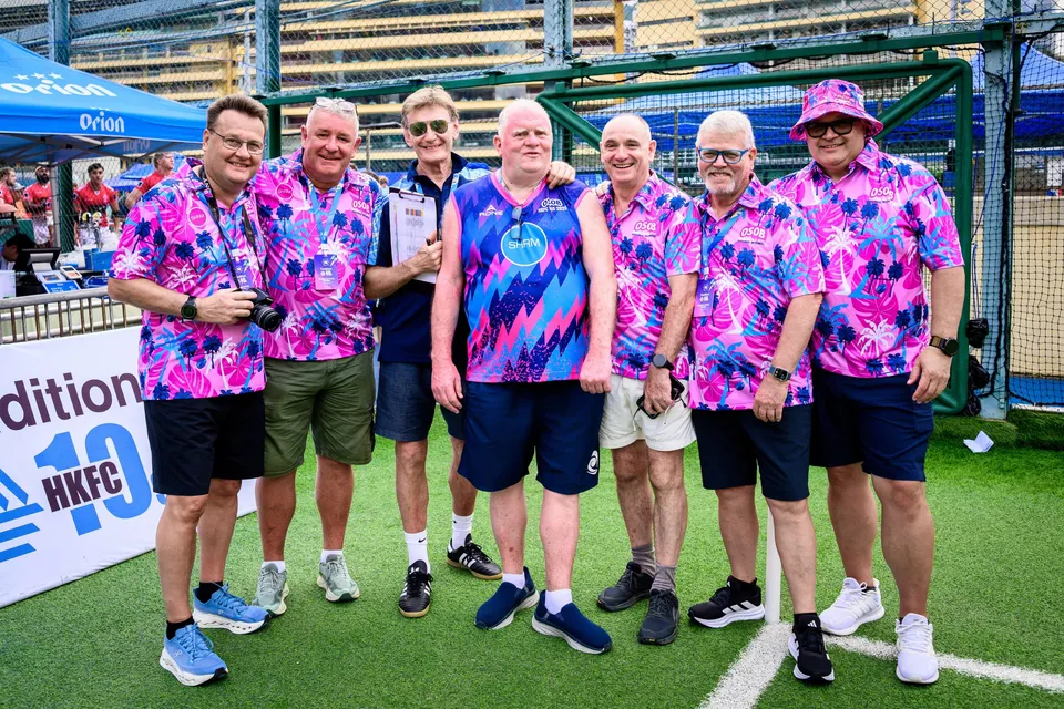 Seven men in pink floral shirts pose on the grass beside an HKFC 10s board, 2026 HKFC 10s