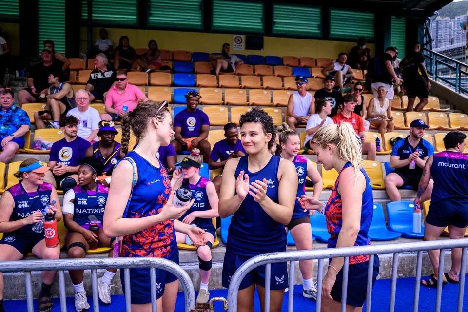 Women in blue and purple kit stand at a barrier below the stand, one applauding, at the 2026 HKFC 10s