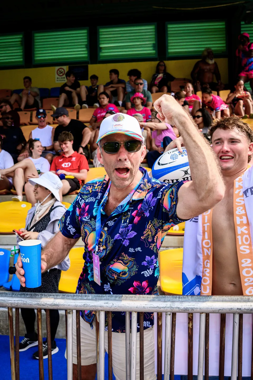Man in sunglasses and a floral shirt raises a fist in the stand, holding a blue cup, at the 2026 HKFC 10s