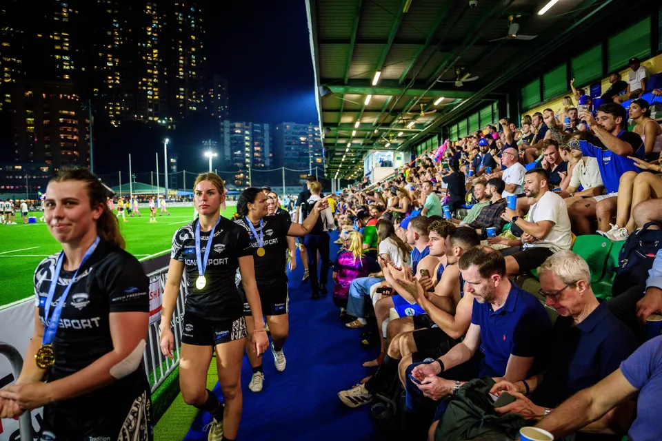Players in black kit with medals walk past a packed stand at night beside the pitch, 2026 HKFC 10s