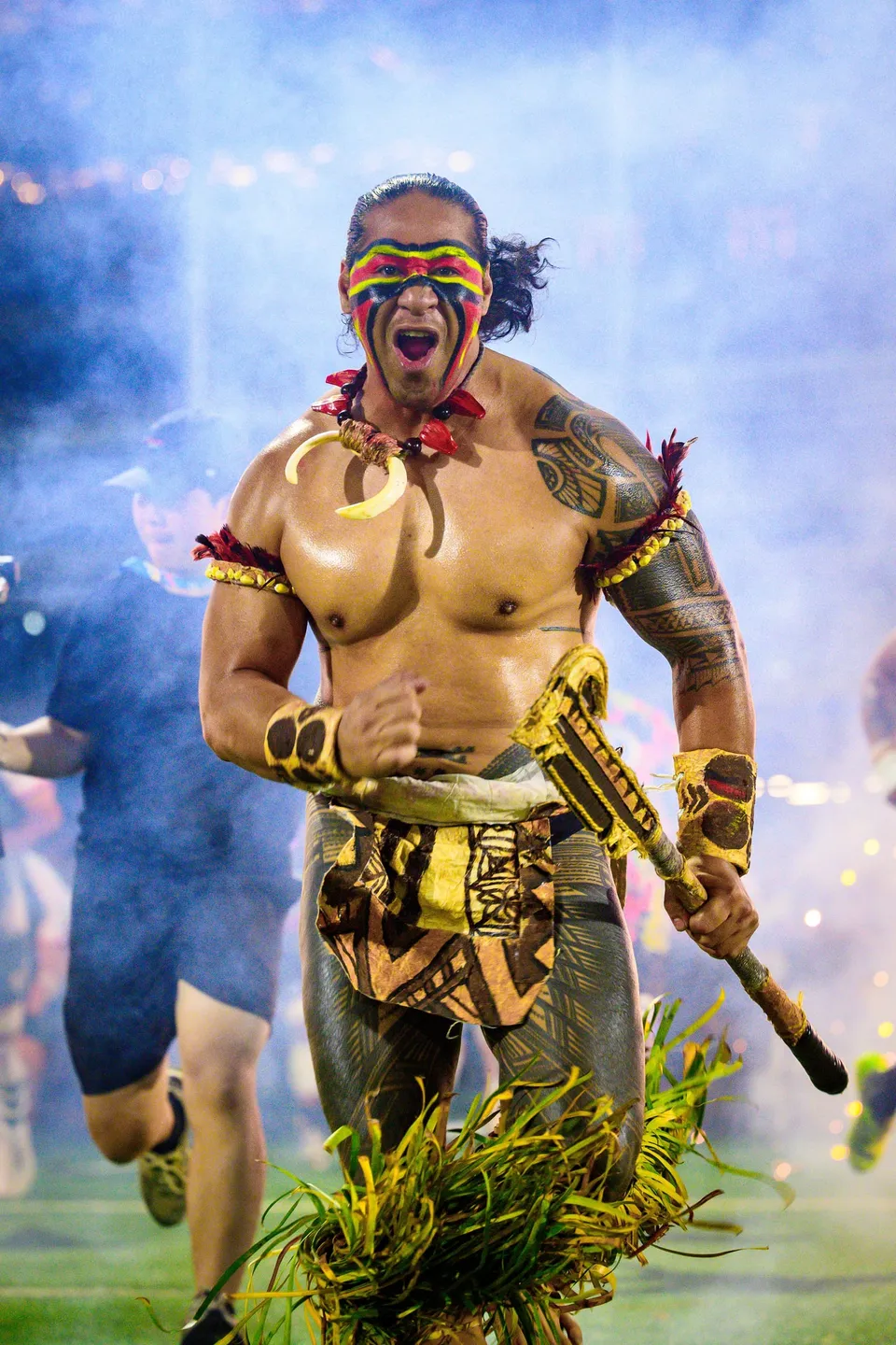 Performer with painted face, feathered armbands and a club walks out through smoke at the 2026 HKFC 10s