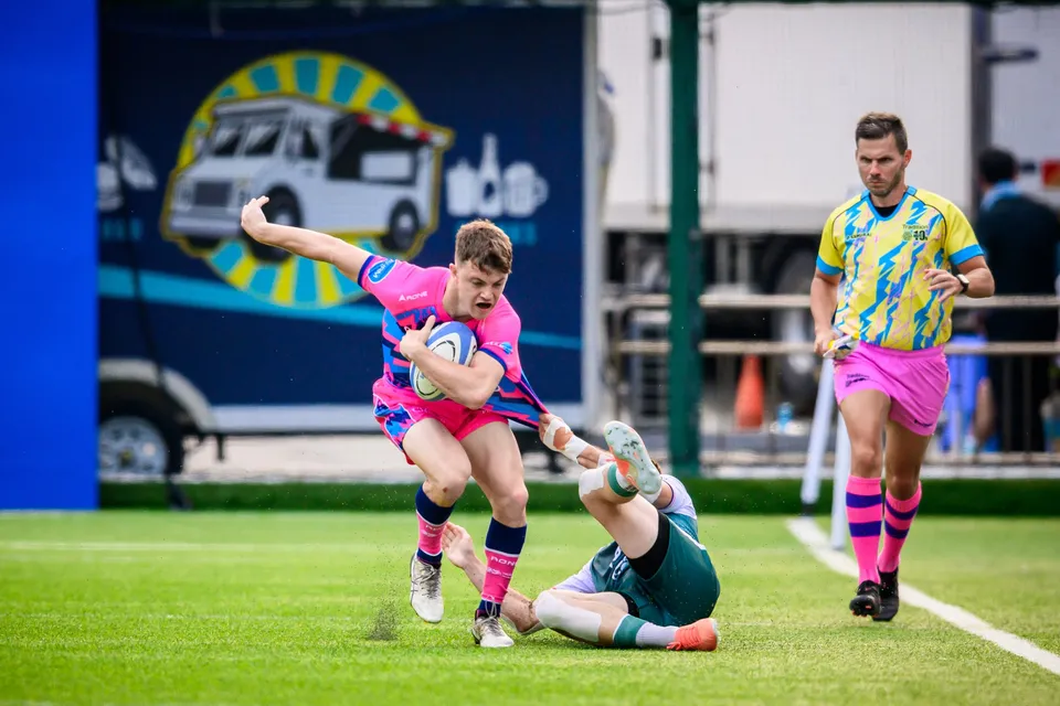 OSOB ball carrier steps away from a Froggies Club tackler sliding on the grass, referee close behind