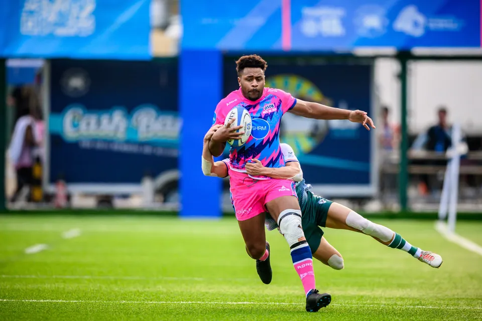 OSOB player in pink and blue carries the ball as a Froggies Club defender dives at his legs