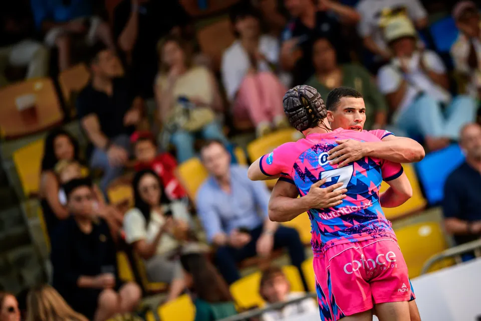 Two OSOB players in pink embrace in front of a stand of spectators with yellow and orange seats