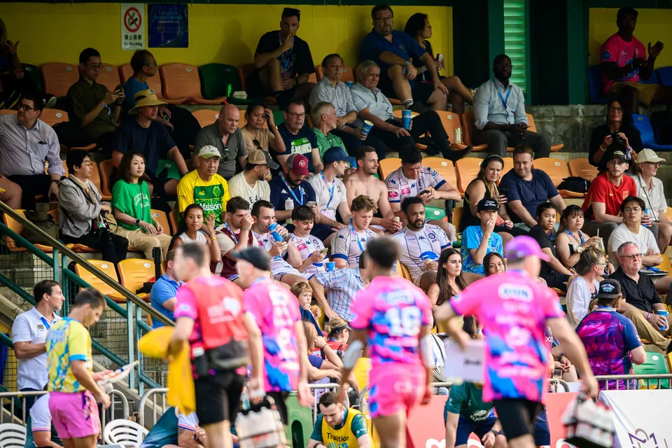 Spectators watch from the stand while two OSOB players in pink walk the touchline and Froggies Club substitutes wait