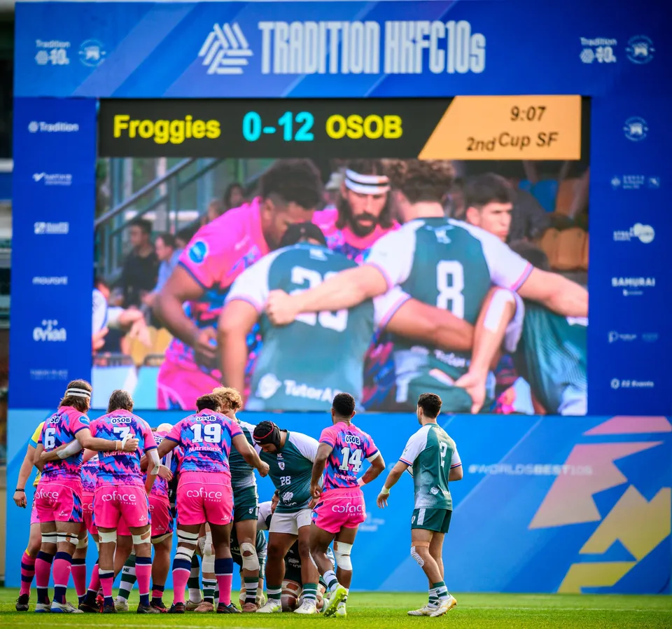 OSOB players in pink gather in a tight huddle below a big screen showing the scoreboard and a close-up of the huddle