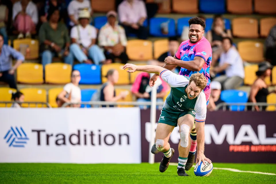 Froggies Club player stumbles forward with the ball as an OSOB defender reaches for him in front of Tradition boards
