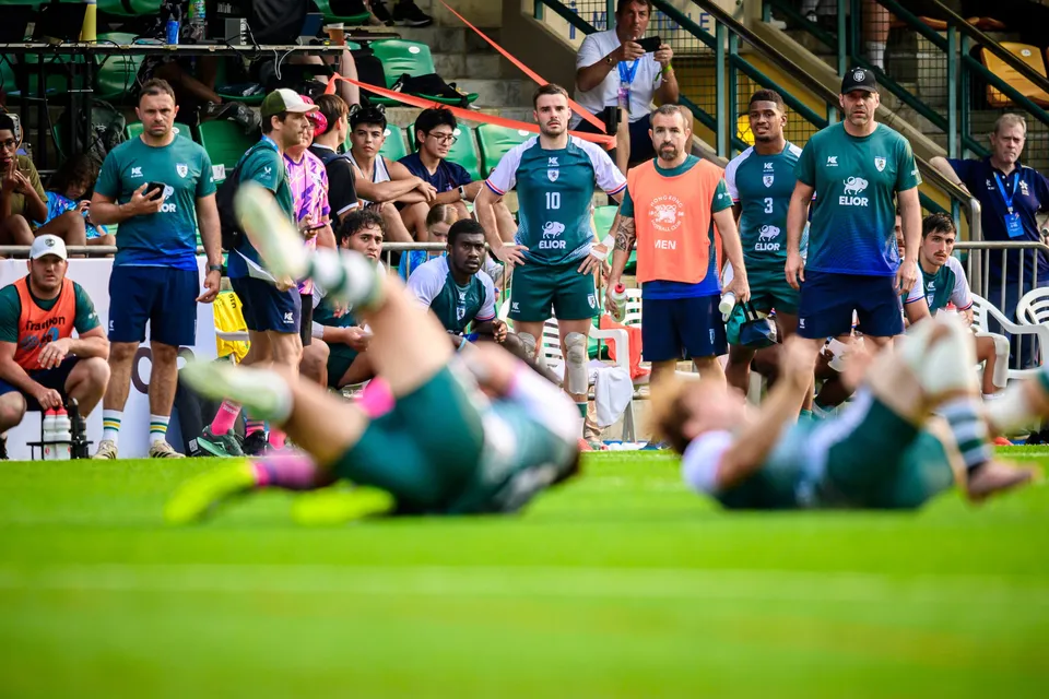 Two Froggies Club players lie on the grass in blurred foreground while teammates and staff stand behind the touchline
