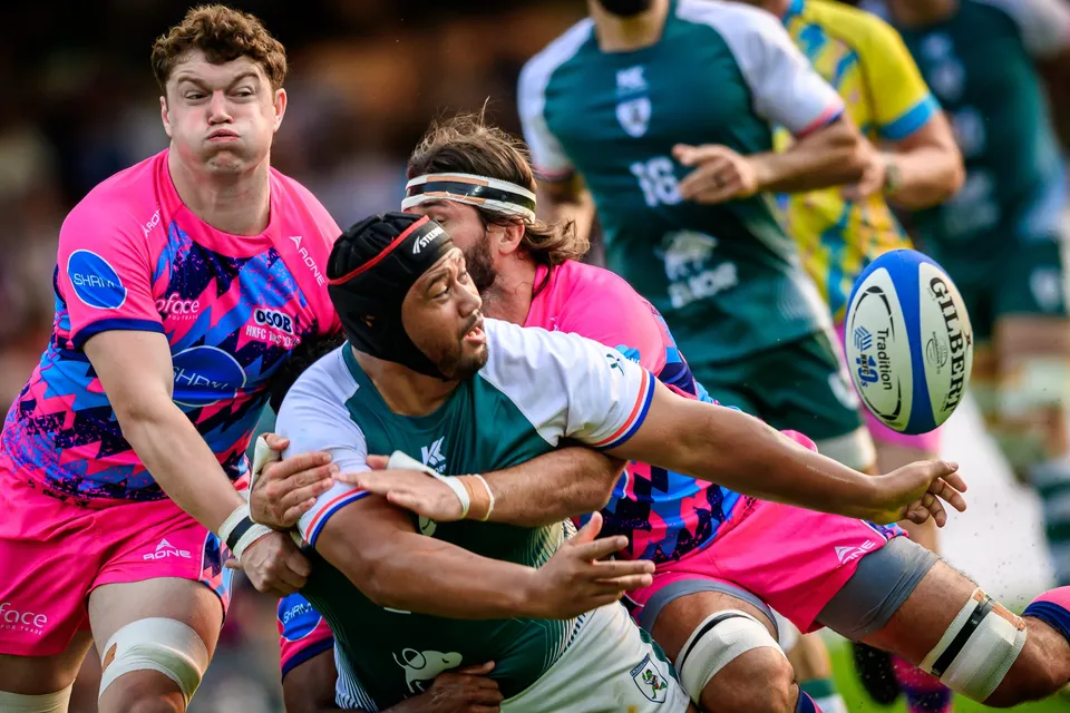 Froggies Club player in a scrum cap passes out of the tackle with the ball in mid-air, an OSOB player grimacing behind
