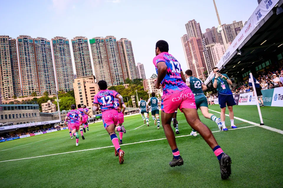 OSOB team in pink jogs onto the pitch toward Froggies Club, with Hong Kong apartment towers behind the stand