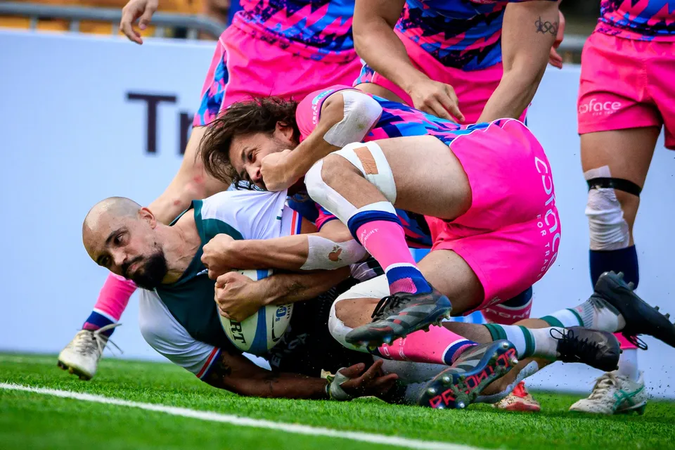 Froggies player in green and white is tackled to ground clutching the ball as OSOB players in pink pile over him