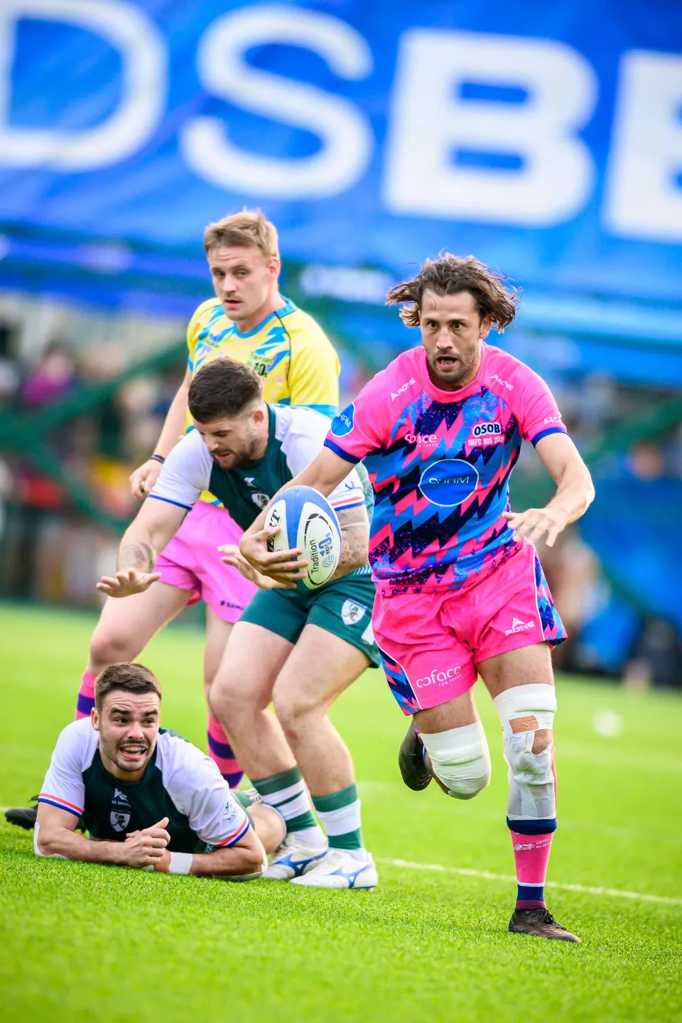 Long-haired OSOB player runs with the ball in both hands past a Froggies player lying on the grass