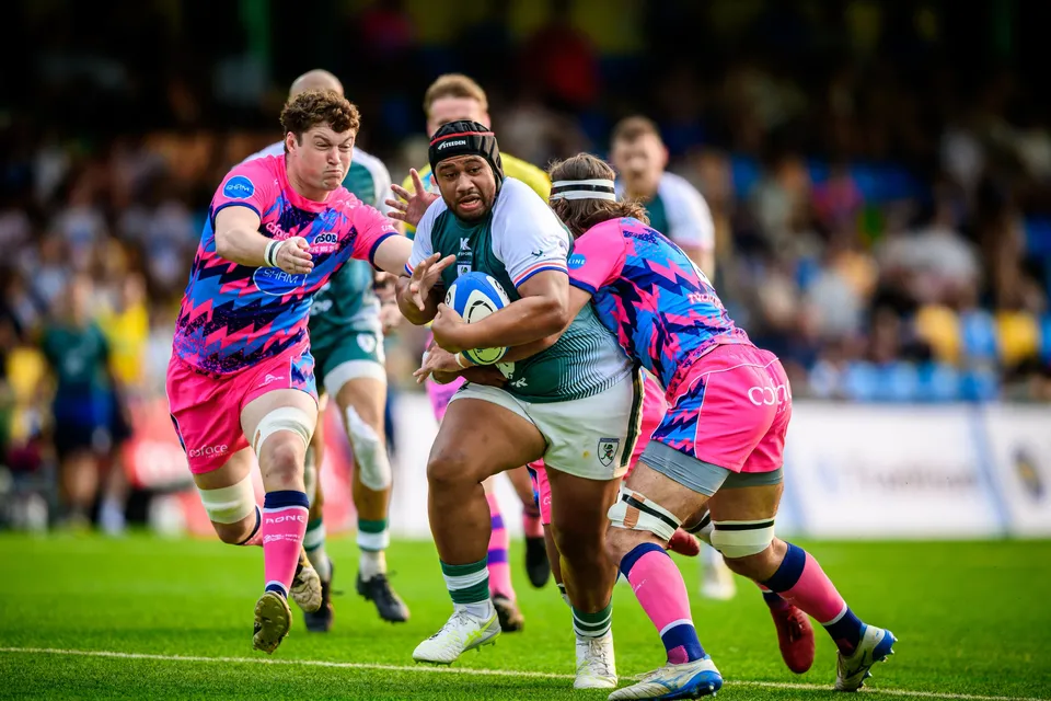 Froggies Club ball carrier in a scrum cap drives through two OSOB defenders in pink, with stands behind
