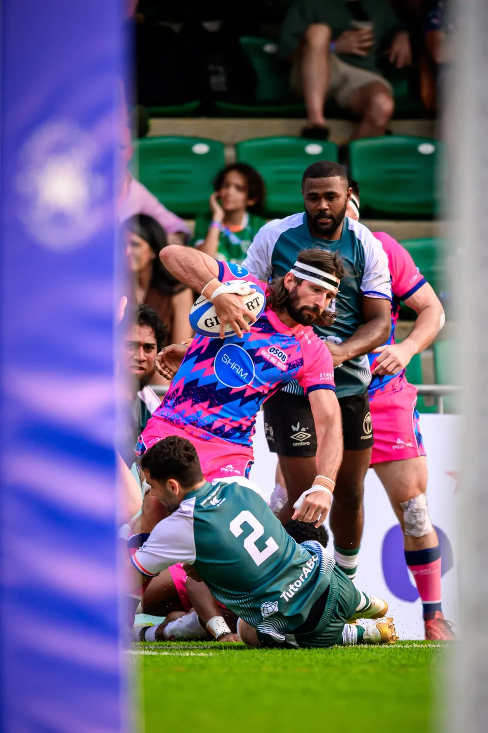 OSOB ball carrier in a headband drives over a fallen Froggies Club number 2 in front of a green-seated stand