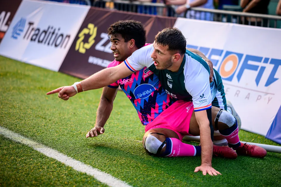 OSOB player points ahead while seated on the grass beside a Froggies Club player, advertising boards behind