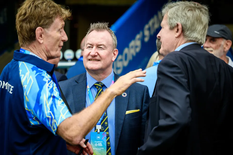 Three men chat in the stand entrance at the 2026 HKFC 10s opening ceremony, one wearing a lanyard and tie