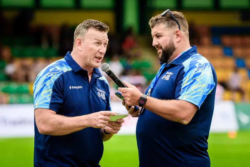 Two men in navy club polos share a microphone for an interview beside the pitch at the 2026 HKFC 10s