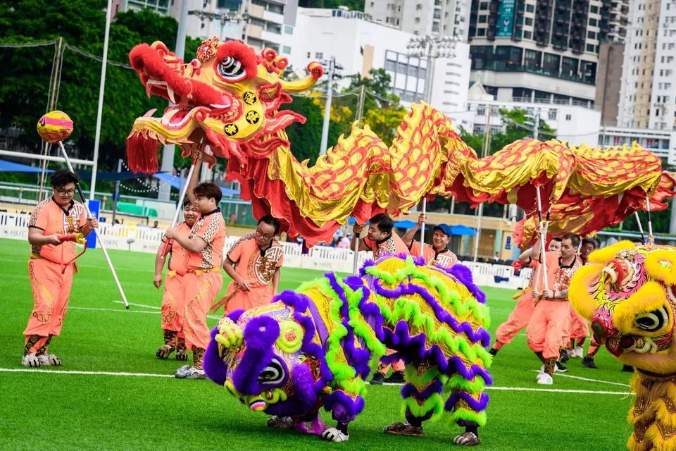 Lion dancers in orange costumes with a purple and green lion head on the grass, 2026 HKFC 10s opening ceremony