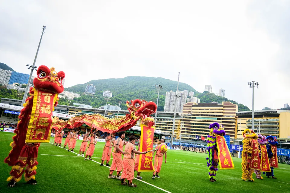 Dragon and lion dancers in orange costumes move across the pitch with hills behind, 2026 HKFC 10s opening ceremony