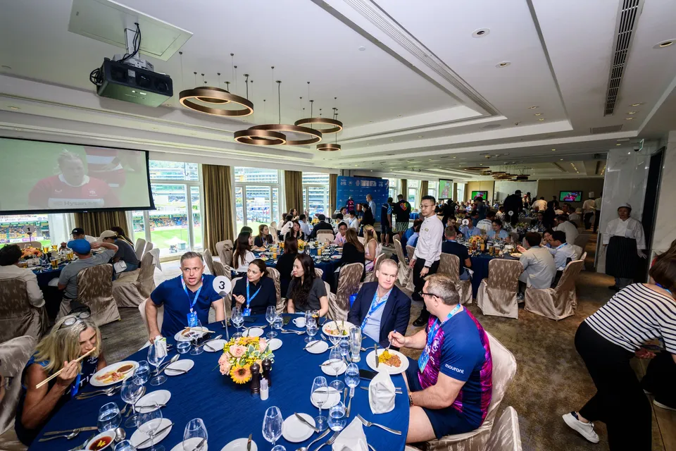 Lunch guests seated at a table with flowers and plates while a screen shows rugby, Patrons' Lunch, 2026 HKFC 10s