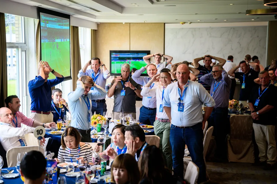 Standing lunch guests with hands on their heads watch a television screen, Patrons' Lunch at the 2026 HKFC 10s