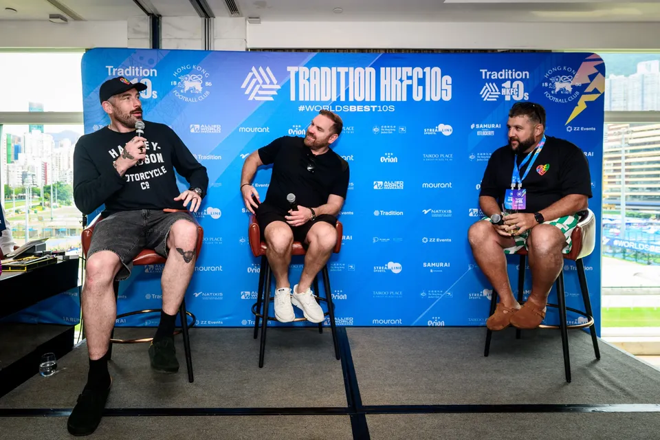 Three men sit on stools with microphones in front of the HKFC 10s sponsor backdrop at the 2026 event