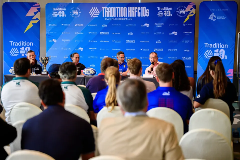 Rows of white chairs face a press panel with a trophy, in front of blue Tradition HKFC 10s banners, 2026