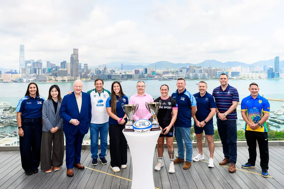 Twelve people stand with two trophies and a rugby ball on a terrace above the harbour, 2026 HKFC 10s