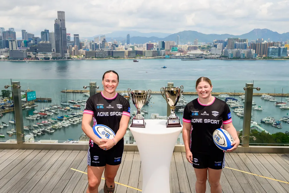 Two women in black team shirts each hold a rugby ball beside two trophies on a harbour terrace, 2026 HKFC 10s