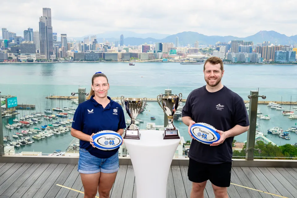 A woman in navy and a man in a dark T-shirt hold Tradition rugby balls beside two trophies, 2026 HKFC 10s