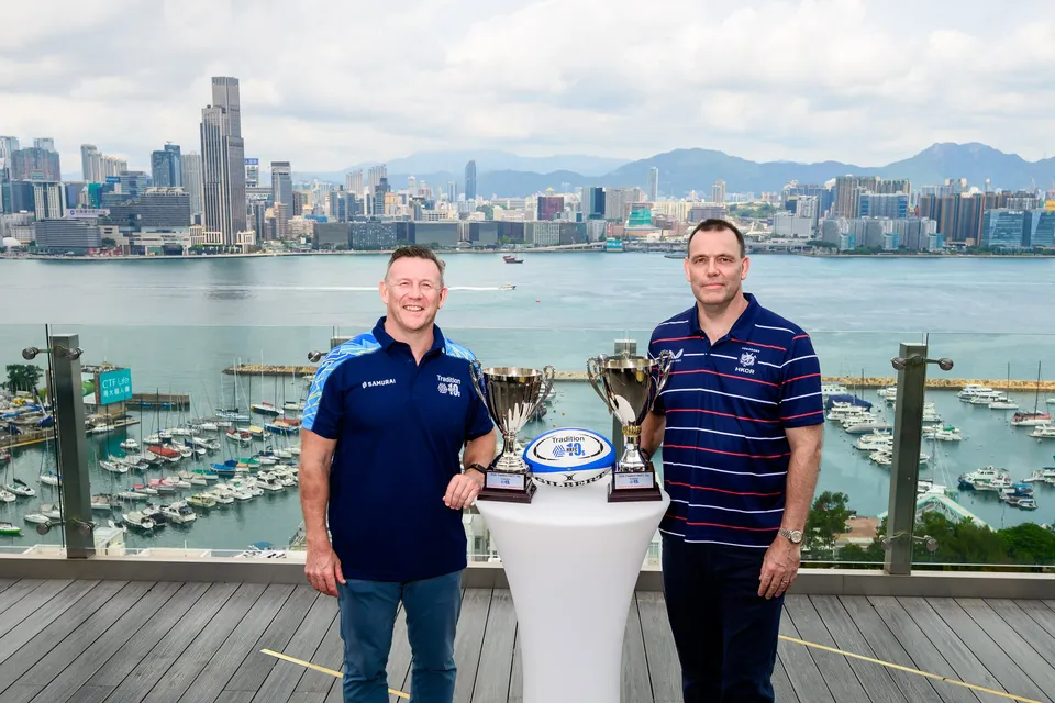 Two men in navy tops stand beside two trophies and a rugby ball with the Hong Kong harbour behind, 2026 HKFC 10s