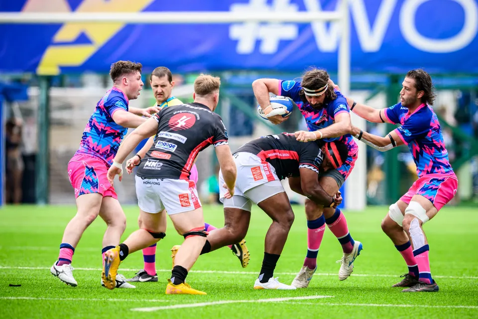 Ball carrier in OSOB kit is held up by two black-shirted opponents as team-mates bind in, a blue banner behind