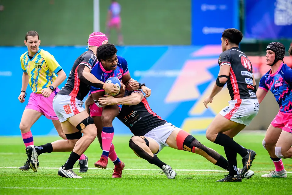 Contested tackle as a referee in yellow and blue looks on and an OSOB player clutches the ball among defenders