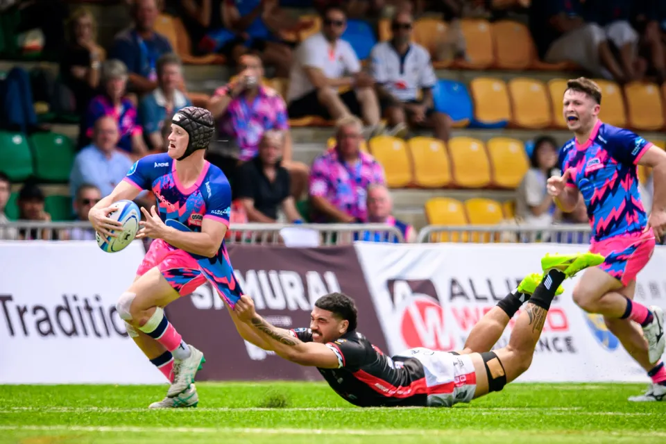 OSOB player in a scrum cap sprints with the ball as a black-shirted opponent dives at his heels, Samurai banner behind