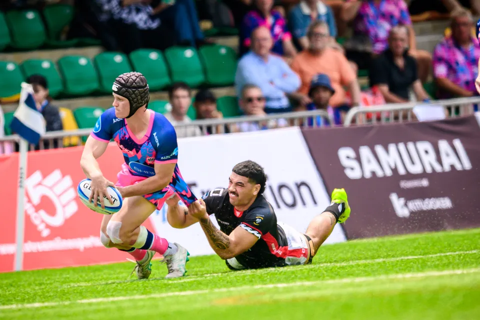 OSOB player cradles the ball in both hands as a diving opponent in black reaches for his feet, spectators in the stand