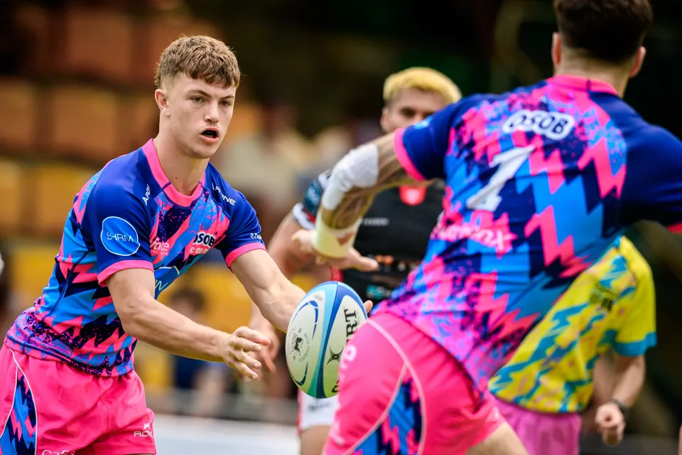 Young OSOB player in a blue patterned shirt looks to pass the ball at close range, a black-shirted opponent behind him