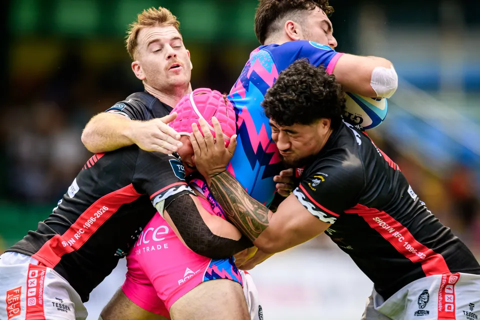 Close-up of an OSOB ball carrier in a pink scrum cap gripped from three sides by black-shirted opponents