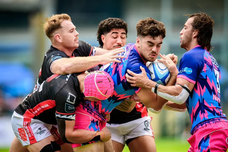 OSOB ball carrier is wrapped up by black-shirted defenders as a team-mate in pink headgear joins the contact
