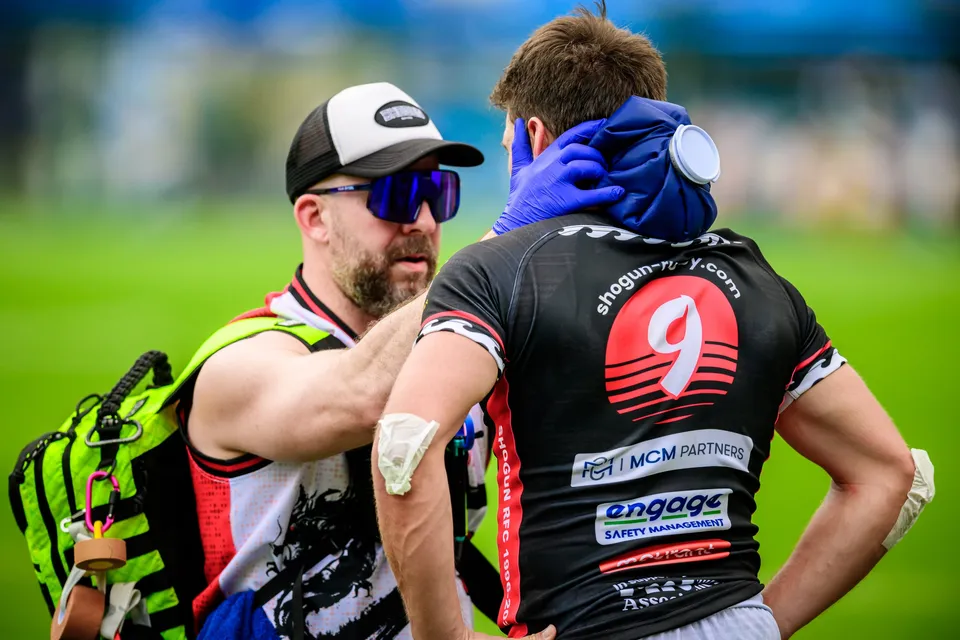 Physio in a cap and sunglasses holds a blue cold pack to the neck of a standing black-shirted number 9