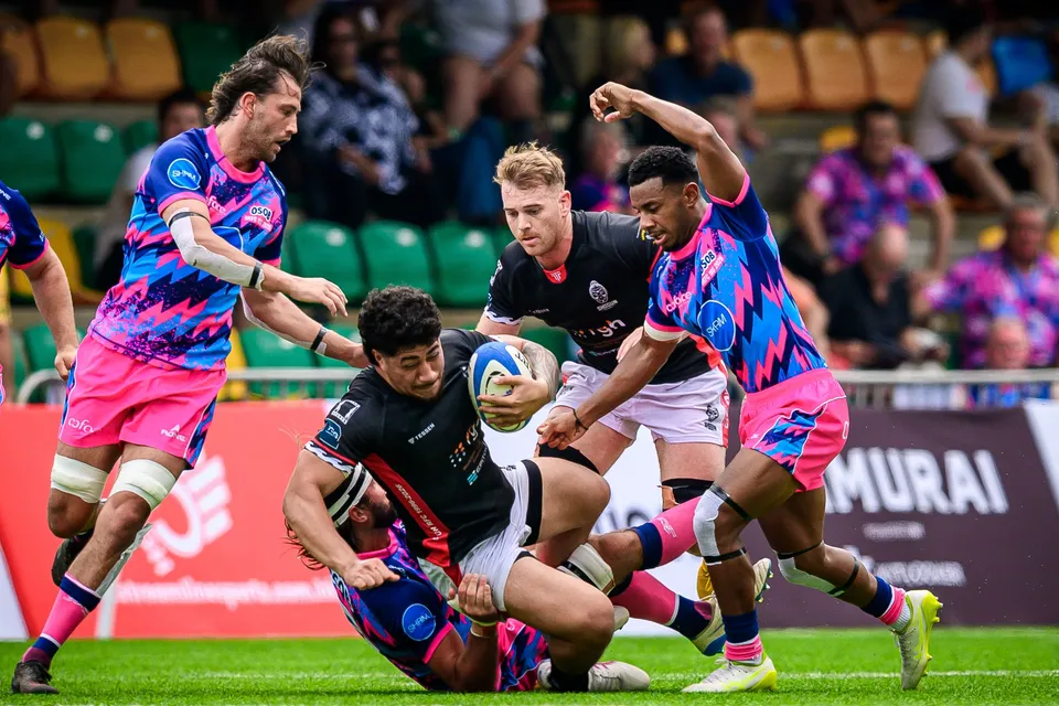 Black-shirted ball carrier is brought down by OSOB defenders near a Samurai banner while a team-mate raises an arm