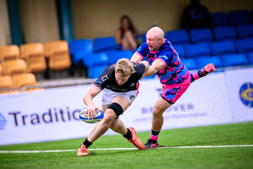 Black-shirted player carries the ball low and fast past Tradition boards while a bald OSOB defender chases from behind